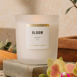 Lit, white soy candle in frosted vessel.  "Bloom" is in black on label with "limited edition" in gold. Warm-coloured stone background with pink and yellow orchid peeking behind. 