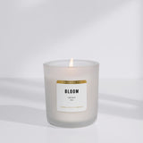 Lit, white soy candle in frosted vessel. "Bloom" is in black on label with "limited edition" in gold. White background.